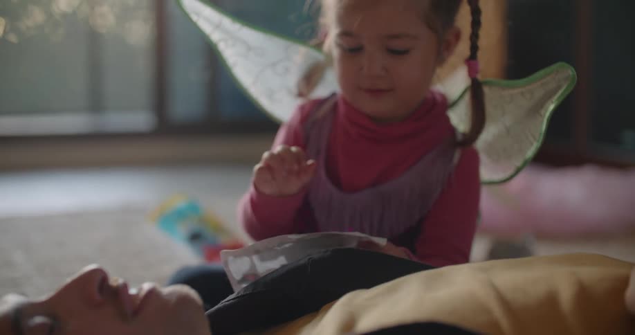 A man lies on the floor of a living room while a young girl attaches stickers to his face