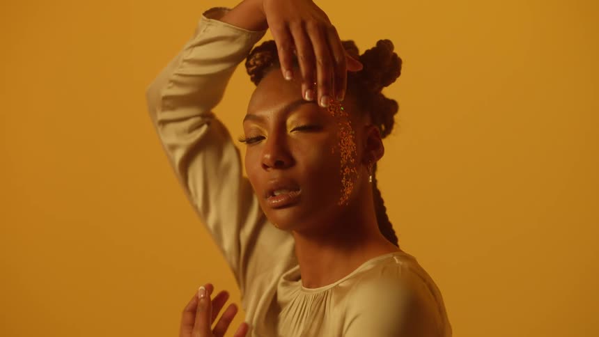 A model runs her hand along her cheek while posing in a studio with yellow walls