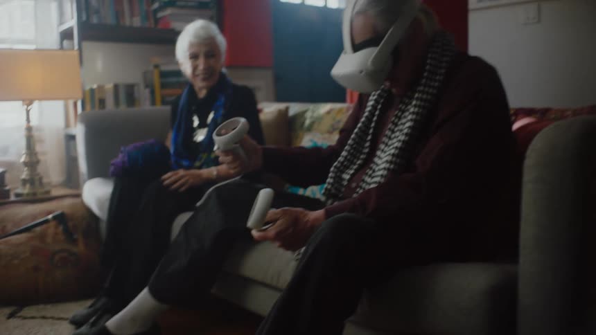 Woman sits on couch smiling as she watches her husband use vr goggles