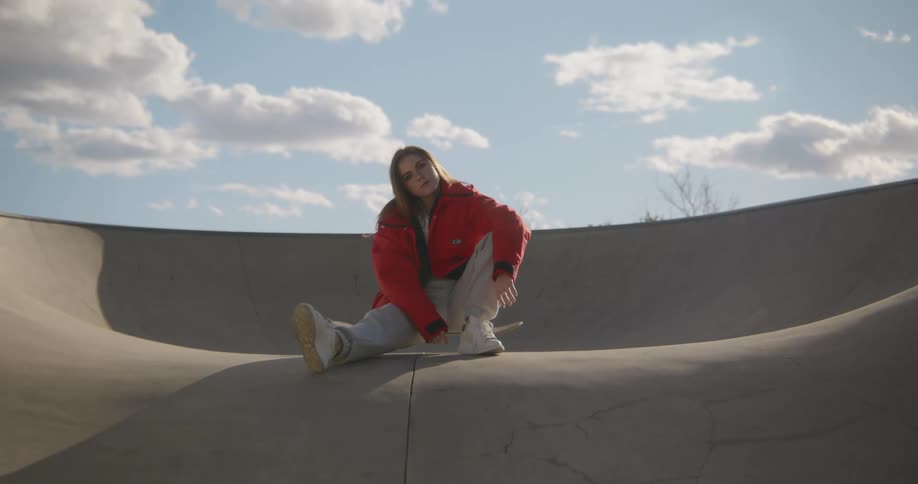 A woman sits idly atop a skateboard in a skate park during the day