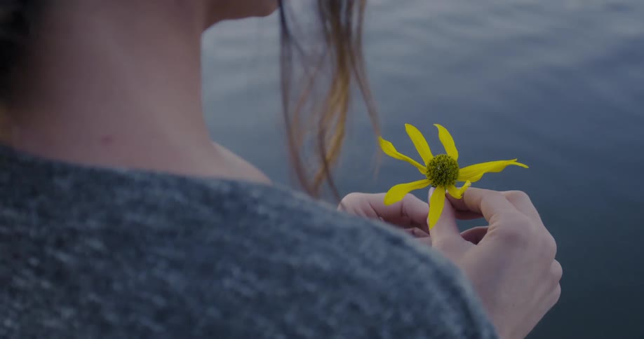 A woman standing twirling a yellow flower in her hands as she looks out at the water