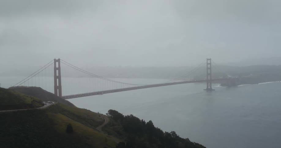 Automobiles travel on a road leading to a suspension bridge stretching across the water and into the fog on the other side