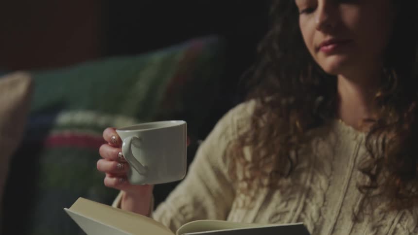 A woman sitting on a couch with a book reaches for a coffee cup sitting on a table and takes a drink