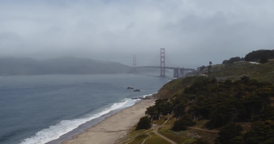 Waves roll over a beach and there is a suspension bridge leading through fog to the other side