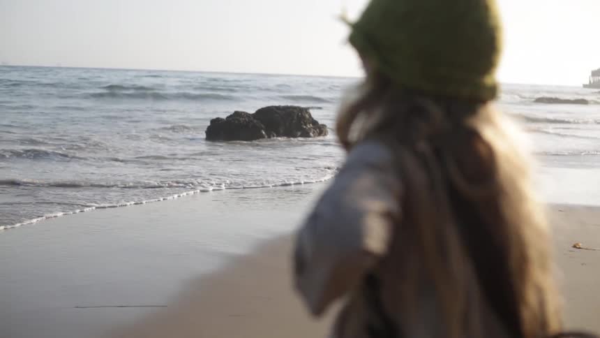 A girl in a puff hat throws something into the ocean