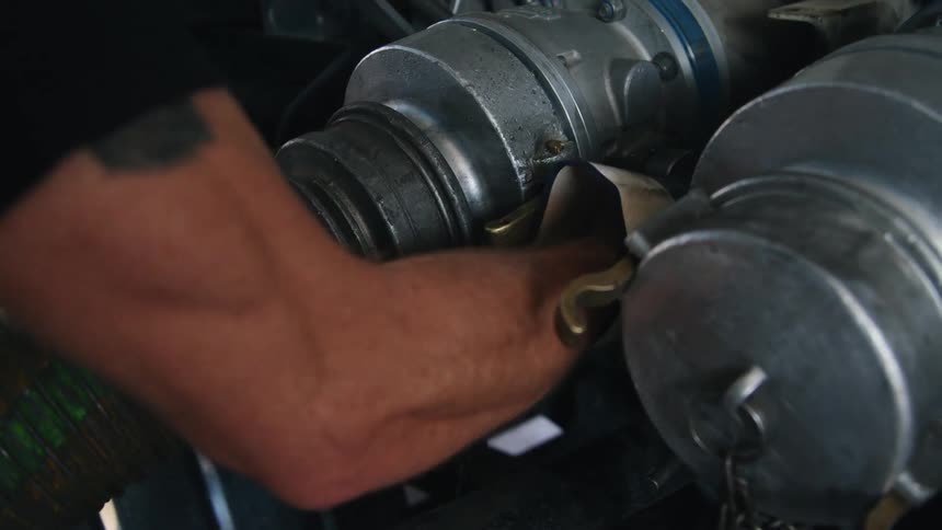 A man with a muscular arm ensures that a large hose is securely connected to the corresponding metal port
