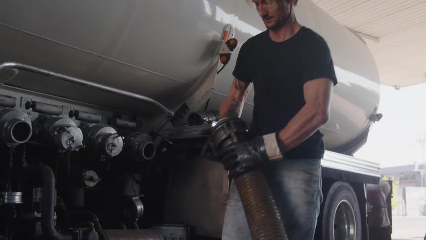 A man wearing gloves puts a piece on a large hose and pulls it near a tanker truck