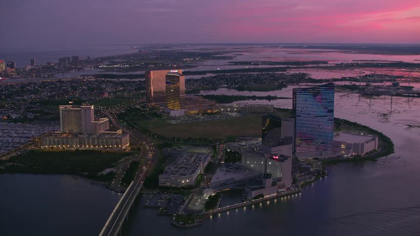 An aerial view of lit up office buildings on the shore with wind turbines in the water and cars driving on a street