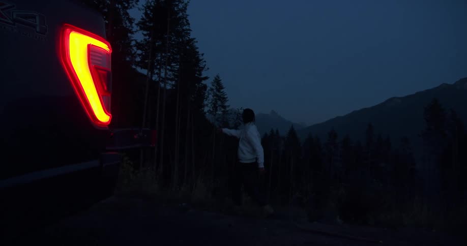 A man stands behind his car at night and angrily throws something far in to the air