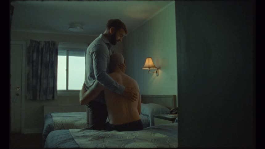 A man sits on a hotel room bed and hugs a man standing next to him as he rubs his back