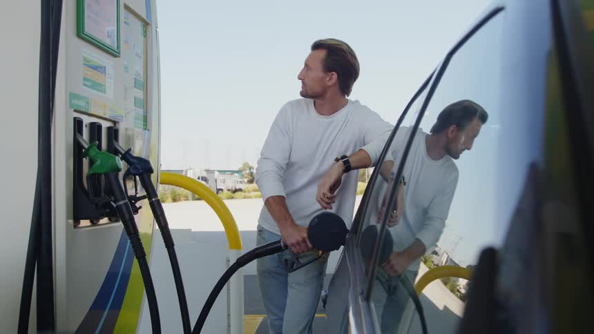 A man is standing next to an automobile as he fills it with gas and puts the gas hose back on the pump