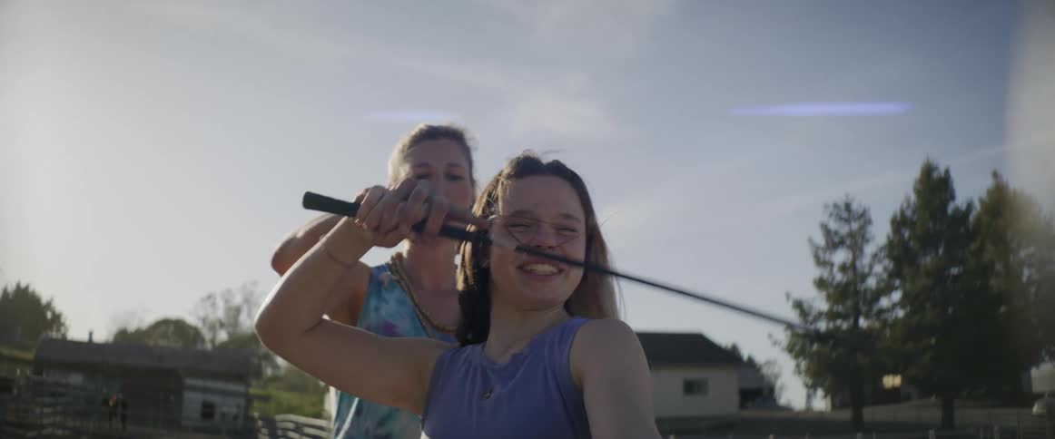 A woman standing behind a teen holding a rod and showing her how to work the lead in a paddock