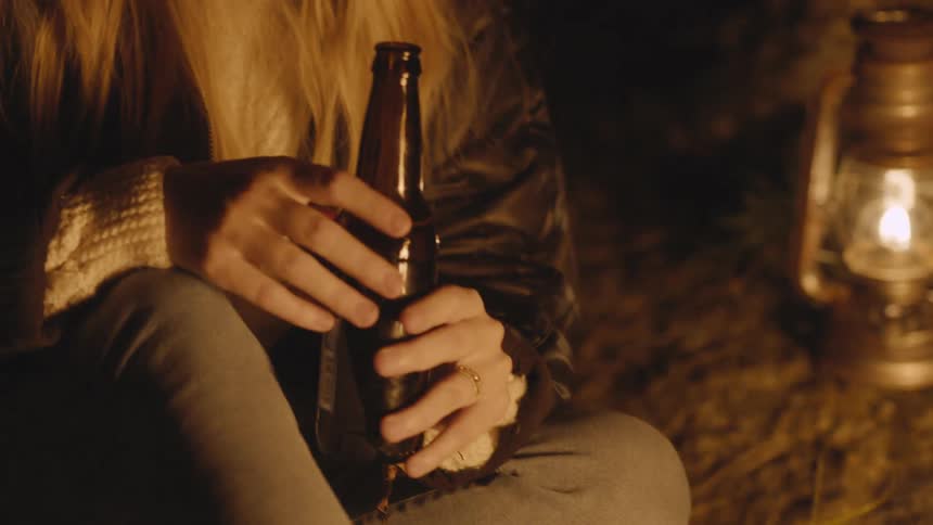 A woman sitting by a campfire holds a beer
