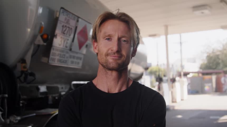 A man is standing at a gas station looking ahead and he wipes his brow