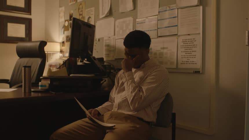 An upset man reading a paper while sitting in an office covers his face with one hand before setting the paper on a nearby desk
