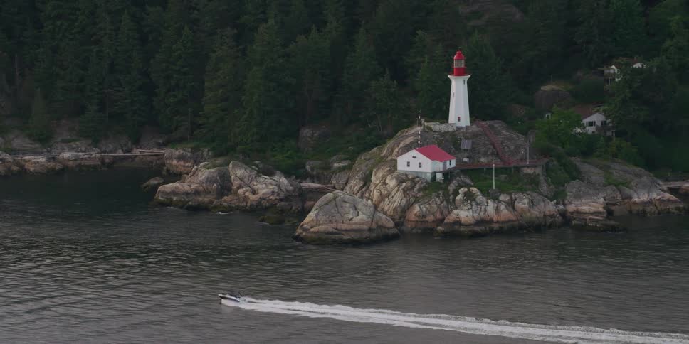 A lighthouse at the foot of a forested mountain overlooks the water as a small boat sails