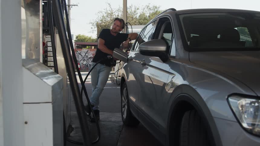 A blonde agitated man in a black shirt pumping gas at a gas station