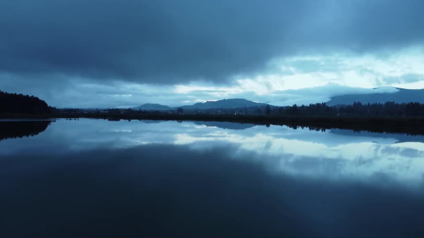 A cloud filled sky reflects on a lake surrounded by forests with mountains in the distance