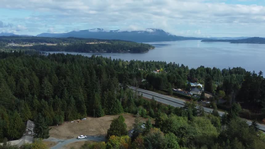 Automobiles travel on a road through a forested rural neighborhood along the coast with mountains in the distance under a cloud filled sky