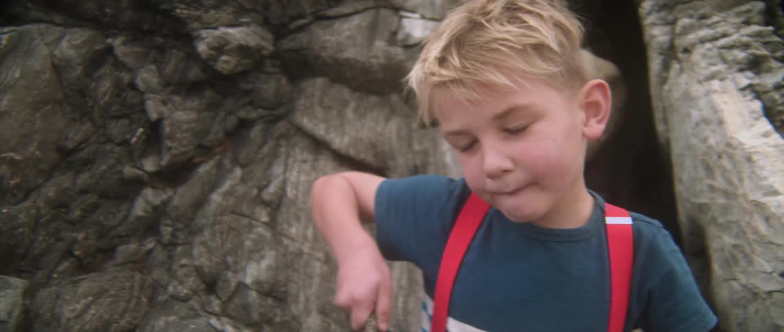 A boy lifts a stick while sitting on rocks