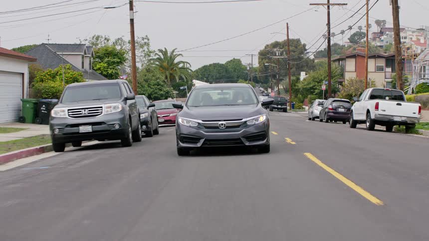 A car with a driver and passenger travels along a road through a residential neighborhood on a cloudy day