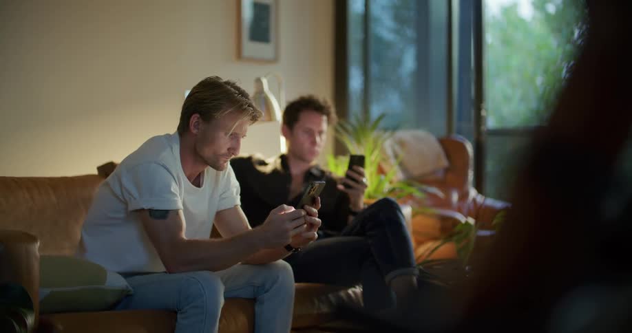 Two men are texting on their phones and sitting indoors