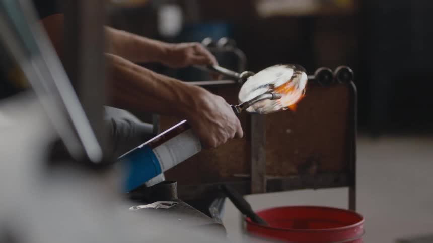 A glass maker torching his piece as he rolls it across a stand