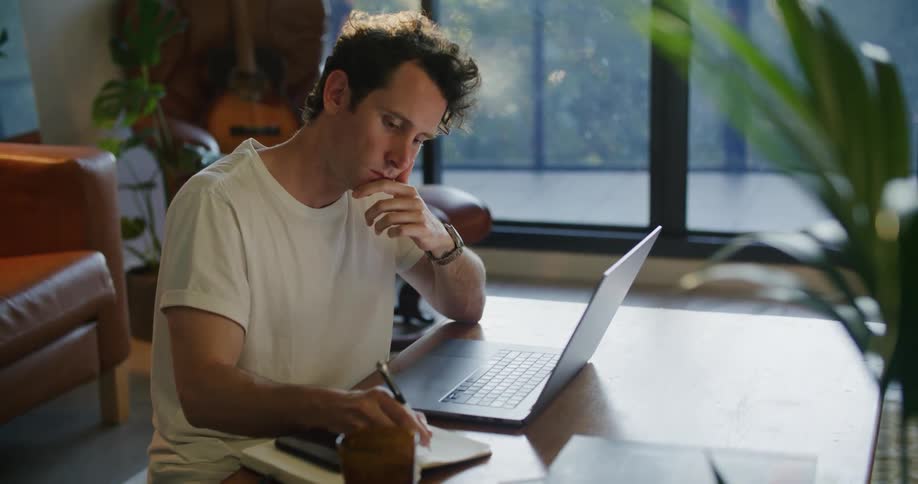 Man sits at coffee table typing on laptop and making notes