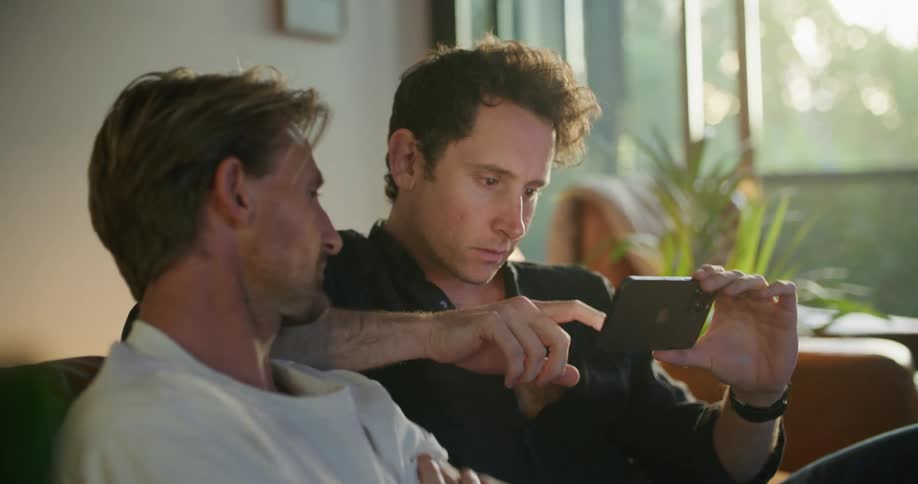 Male friends sit on couch looking over device screen while talking and laughing