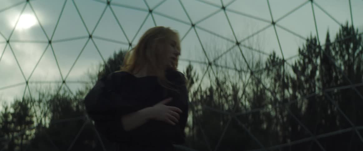 A girl wearing a black dress is outside dancing in front of a fence and there are trees in the distance and the sun is shining through a cloudy sky