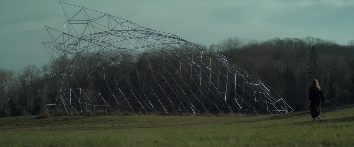 A woman walks through a field towards a tall structure made of bars during the day