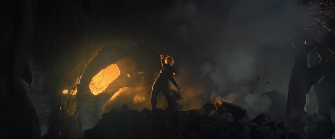 A woman dances in front of sculptures in a dark eerie area with fog rising