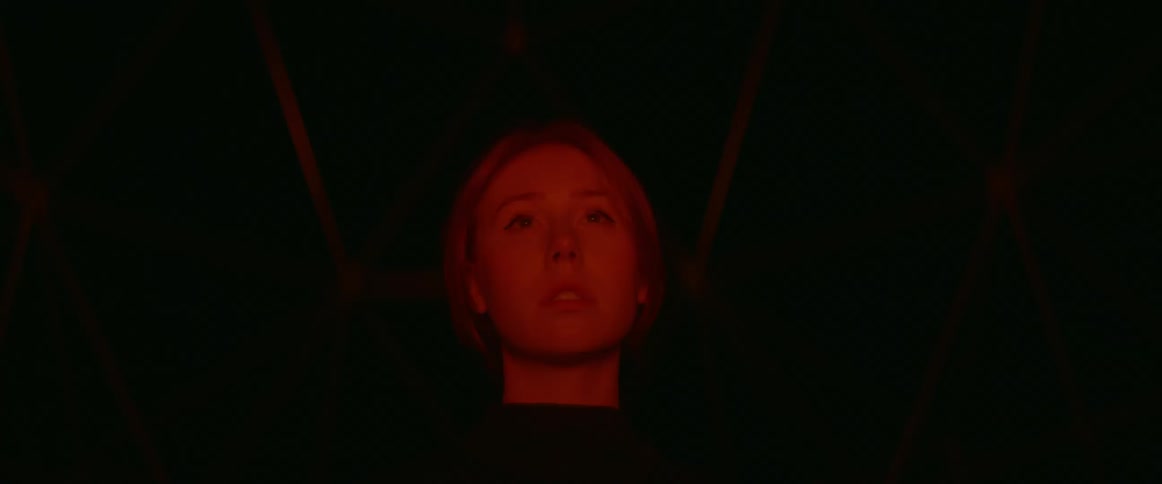 A woman stares fearfully in the distance while standing in a dark structure illuminated by a red light