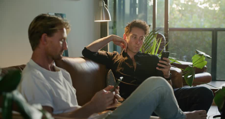 Two men are talking while sitting on a couch and scrolling on cell phones