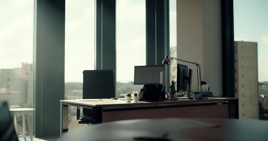 The camera pans to show a computer desk in front of an office window looking out onto a city landscape of tall buildings and also trees