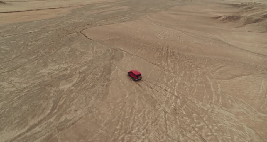 Red jeep drives across sand marked with multiple tire tracks