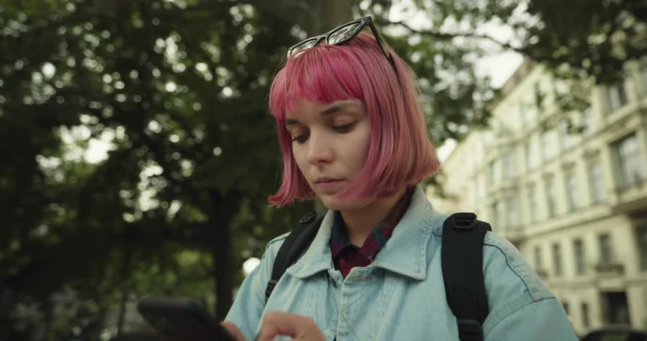 A teenage girl with pink hair and sunglasses on her head is outside near trees using her cell phone