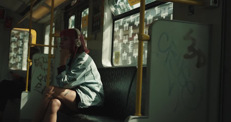 A teenage girl is sitting in a seat on a train with headphones on her head and she looks out the window