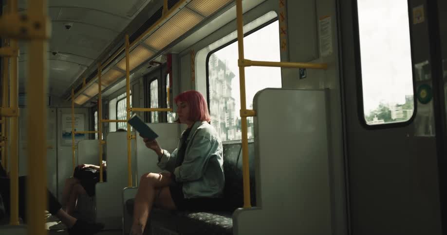 A teenage girl with pink hair is sitting on a train reading a book