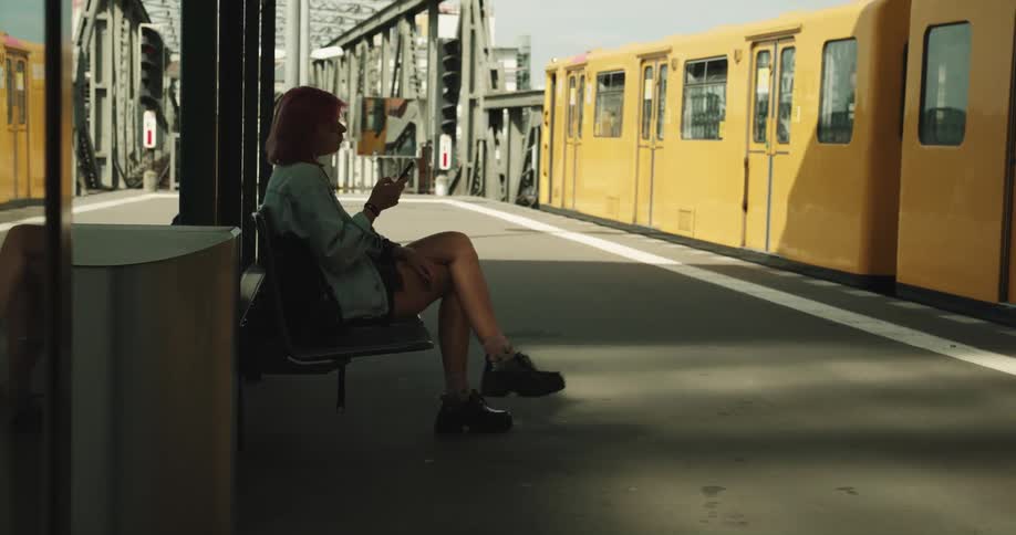 A teenage girl with pink hair is sitting on a bench at a train station using her cell phone