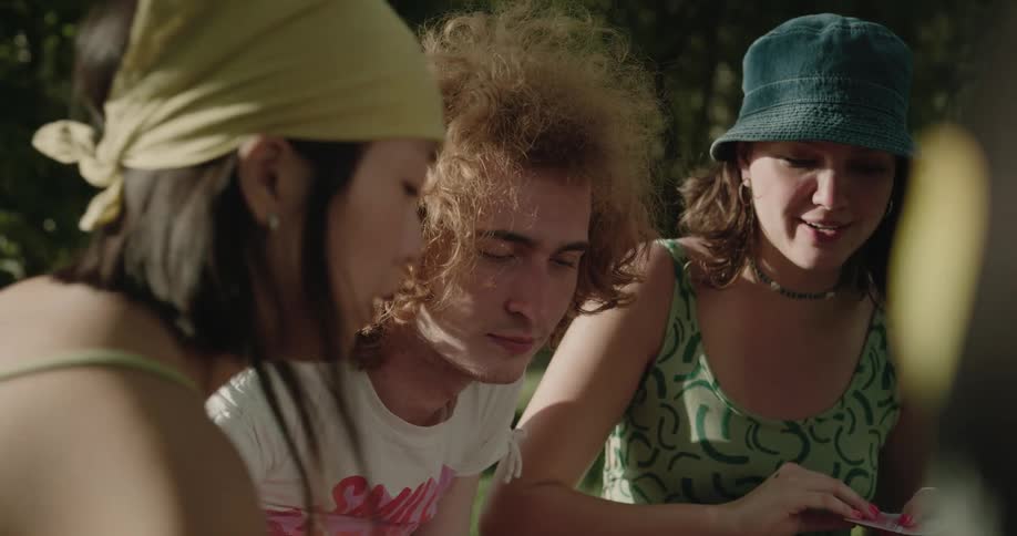 A group of friends are sitting outdoor in the grass and laughing while playing a game of cards