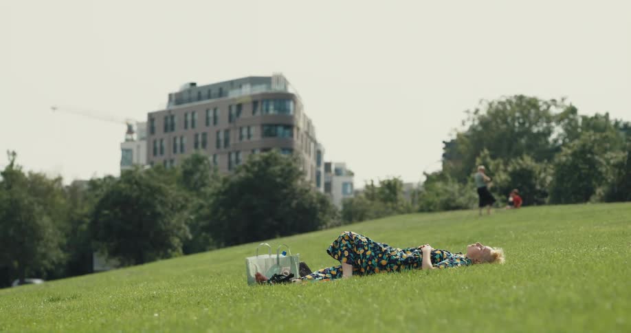 A young woman sitting in field in a public park takes off her shoes and lays in the grass for a moment before sitting back up and retrieving her laptop
