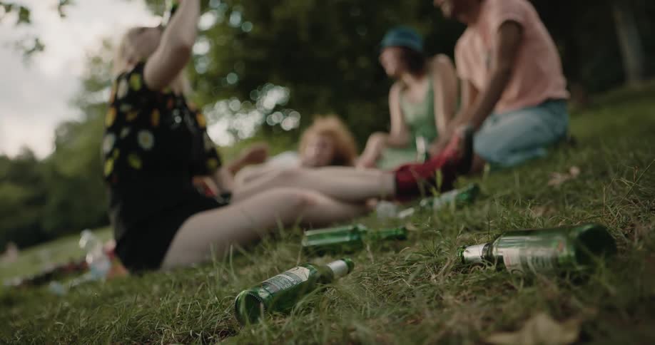 A teenage girl in a park sits with three friends and drinks a beer and throws the bottle to the side where other beer bottles lie