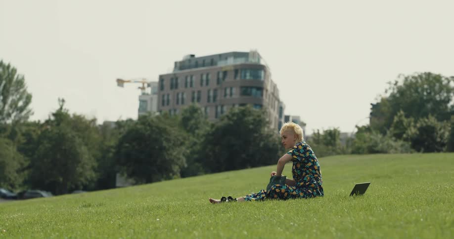 Young short haired blonde woman sitting over the grass on a hill in a park eating a sandwich