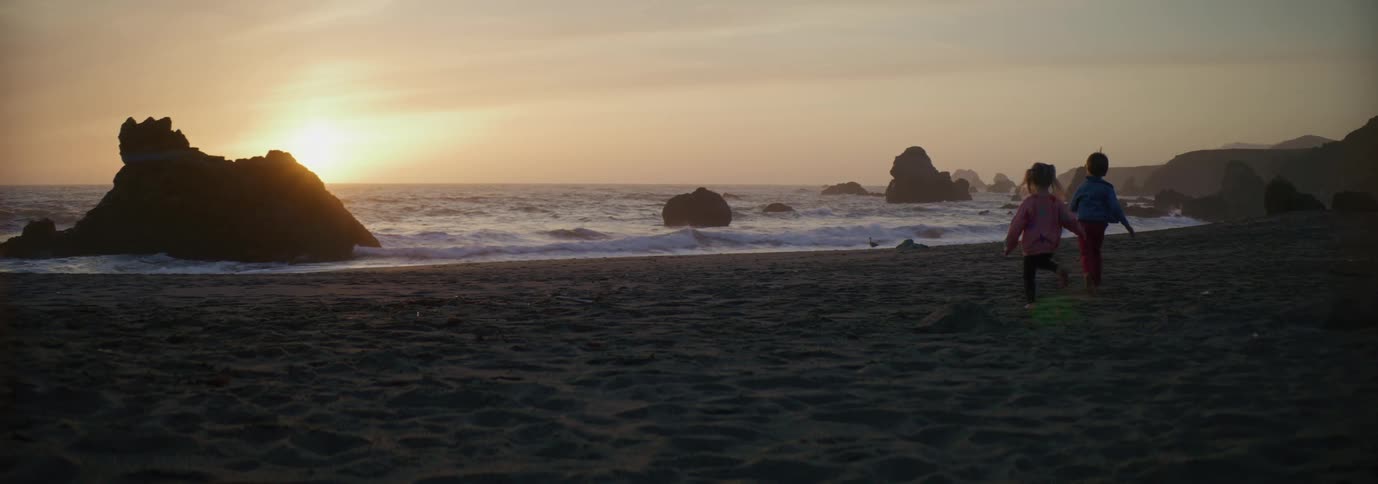Kids run across beach toward surf as sun shines from horizon