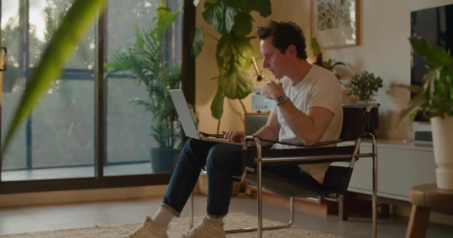 A man is sitting in a chair with a laptop computer having a video conversation with someone and he is talking and laughing