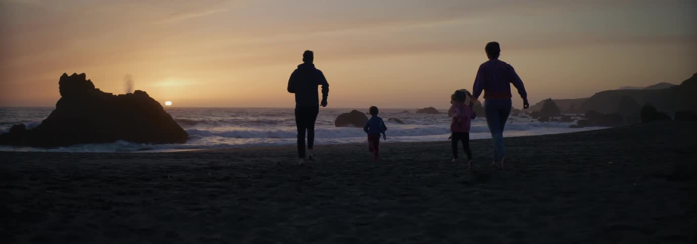 Silhouettes of a family with two small children running across a beach as the sun is setting on the horizon