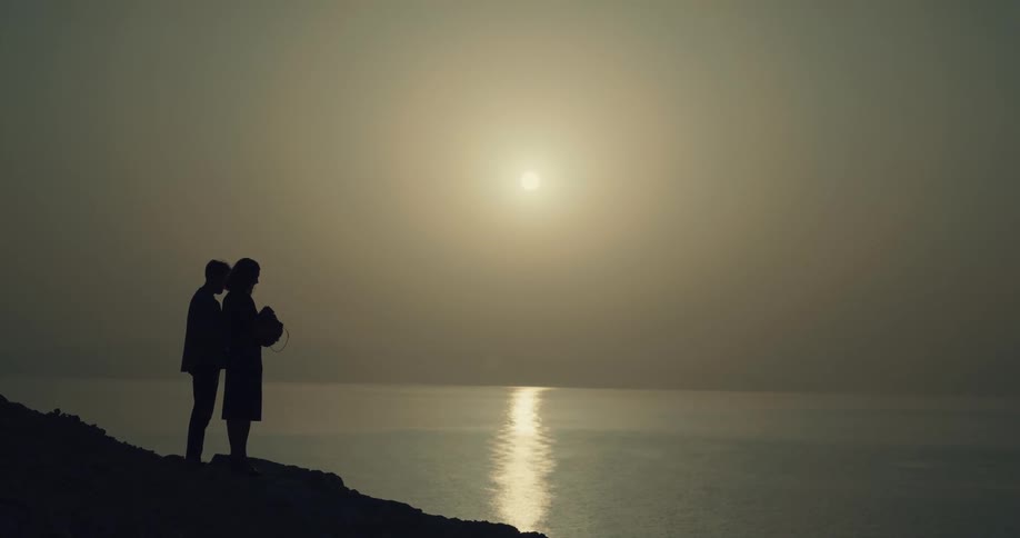 Silhouette of a couple holding an urn with ashes of a loved one near the ocean at sunset