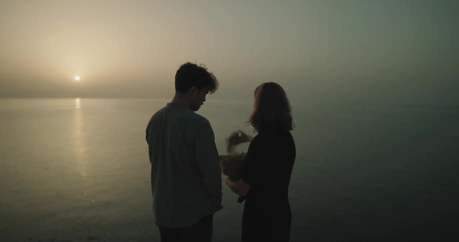 Man and woman are standing near the ocean while the sun is setting