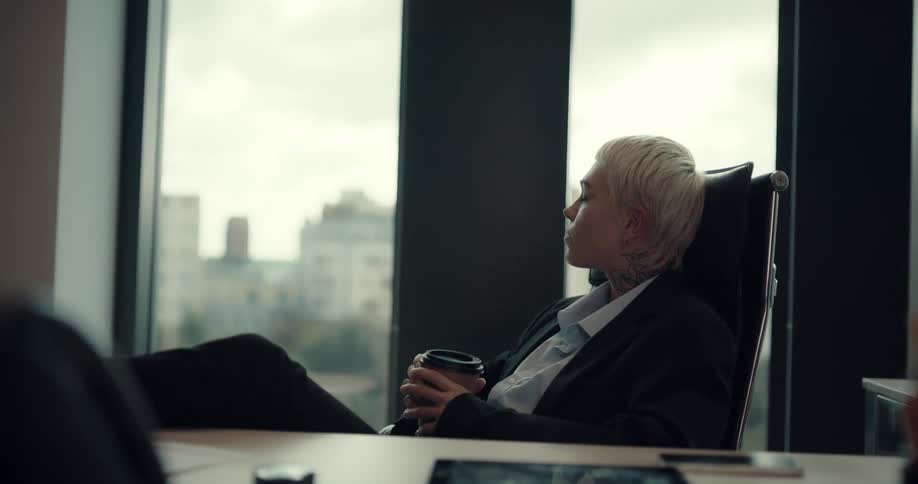 Blonde business woman reclines with feet on desk as she drinks coffee from a to go cup